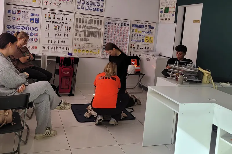 Sala wykładowa OSK w Wołominie: prowadząca w koszulce z napisem „Ratownik” pokazuje resuscytację na fantomie, kursanci obserwują ćwiczenie